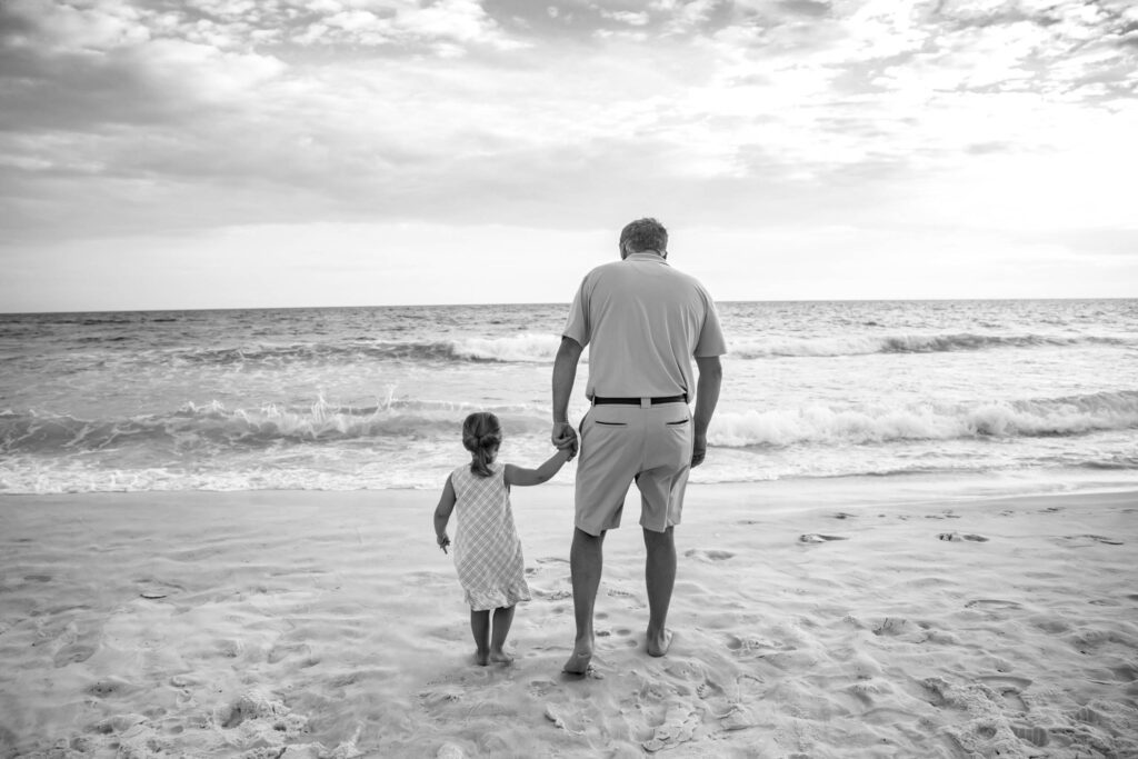 Black and white photo of a father and daughter walking away from the camera on the beach looking at the ocean. This an example of taking a candid photo using composition techniques. This is part of the How to make your family photos and videos look AMAZING blog post. 