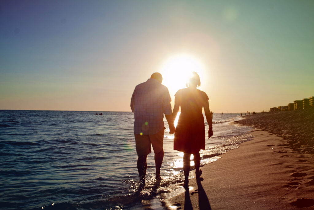 Man and woman, husband and wife, walking on the beach away from the camera at sunset as if walking into the sun. This is an example a candid photo that uses the direct sun to for lighting. This is part of the How to make your family photos and videos look AMAZING blog post. 